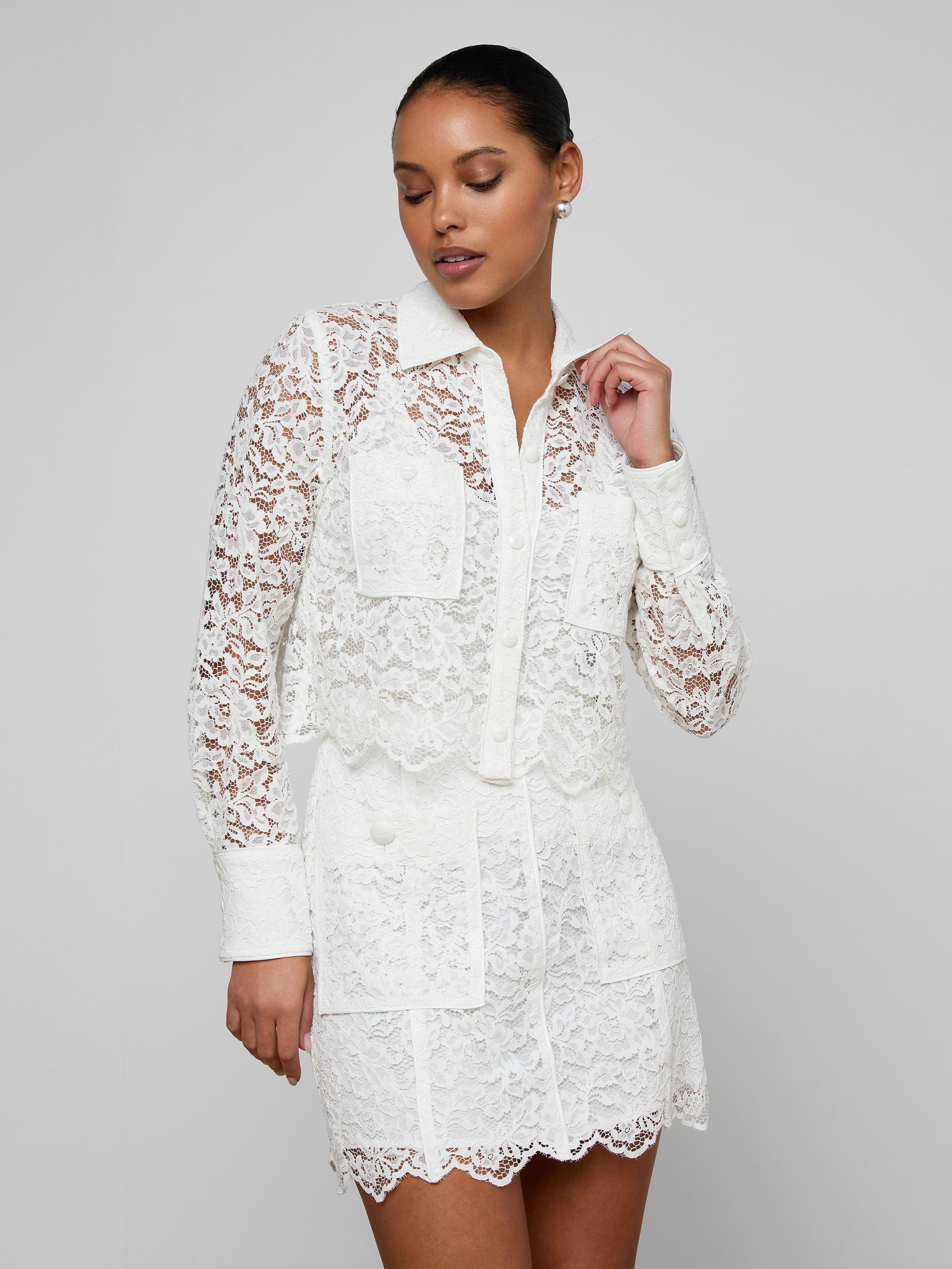 A woman wearing the Roelle Lace Blouse—a white, long-sleeve, floral lace top with patch pockets—stands against a plain light background, looking to the side with one hand raised near her face.