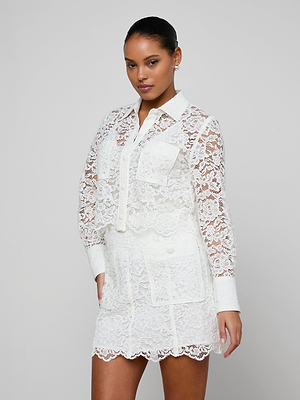 A woman poses in the Roelle Lace Blouse, a white, long-sleeve, semi-sheer floral lace top with a collar, front pockets, and button details against a plain light gray background.