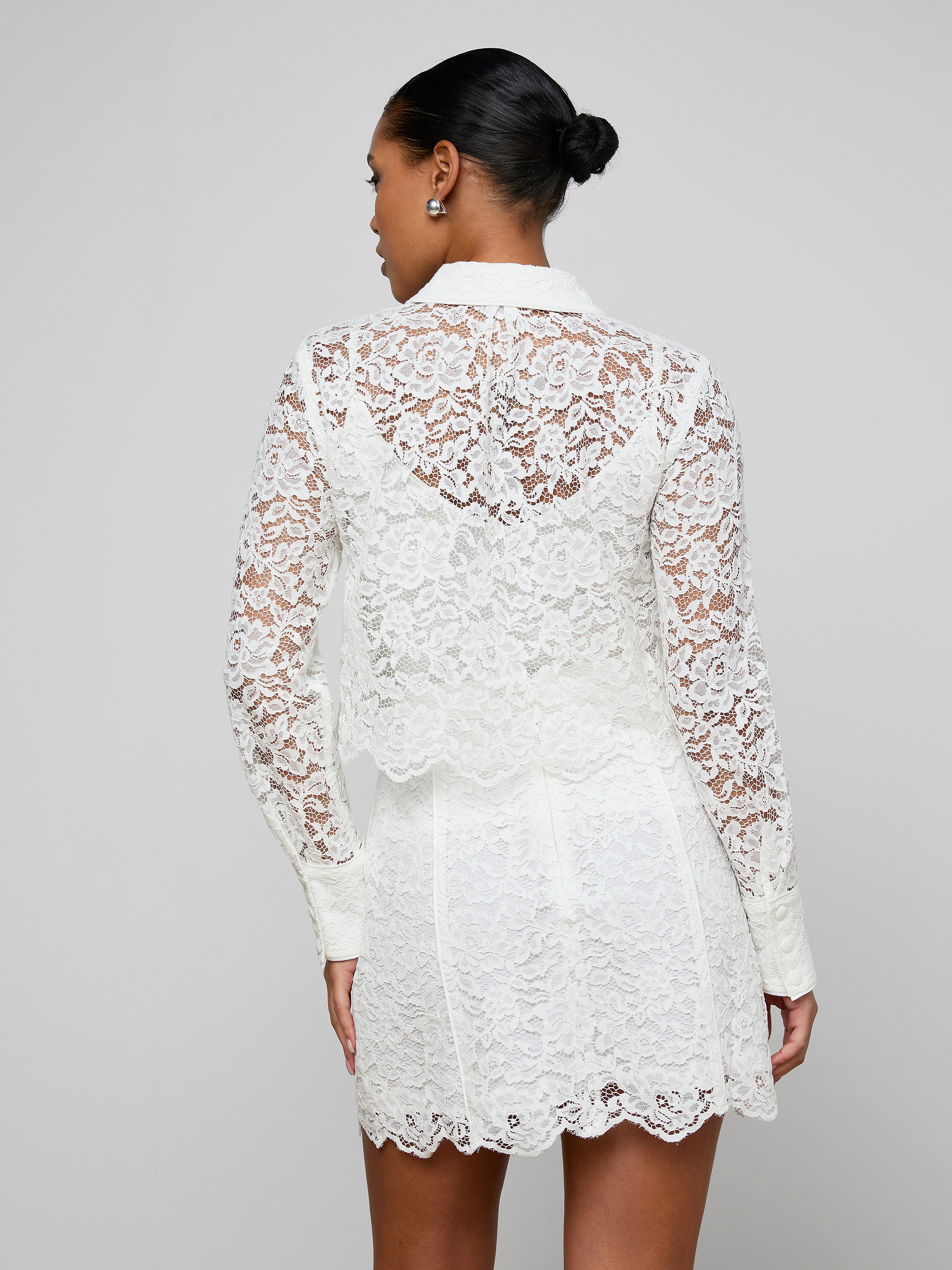 A woman stands with her back to the viewer, wearing the Roelle Lace Blouse—a romantic top made from white floral lace featuring scalloped edges at the hem and collar. Her hair is neatly styled in a bun, set against a simple, light background.