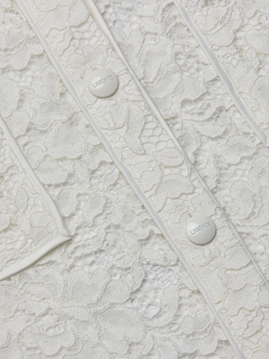 Close-up of the Roelle Lace Blouse showing its white floral lace, decorative stitching, and two white buttons with embossed text. The delicate texture and intricate patterns highlight the blouse’s romantic style.