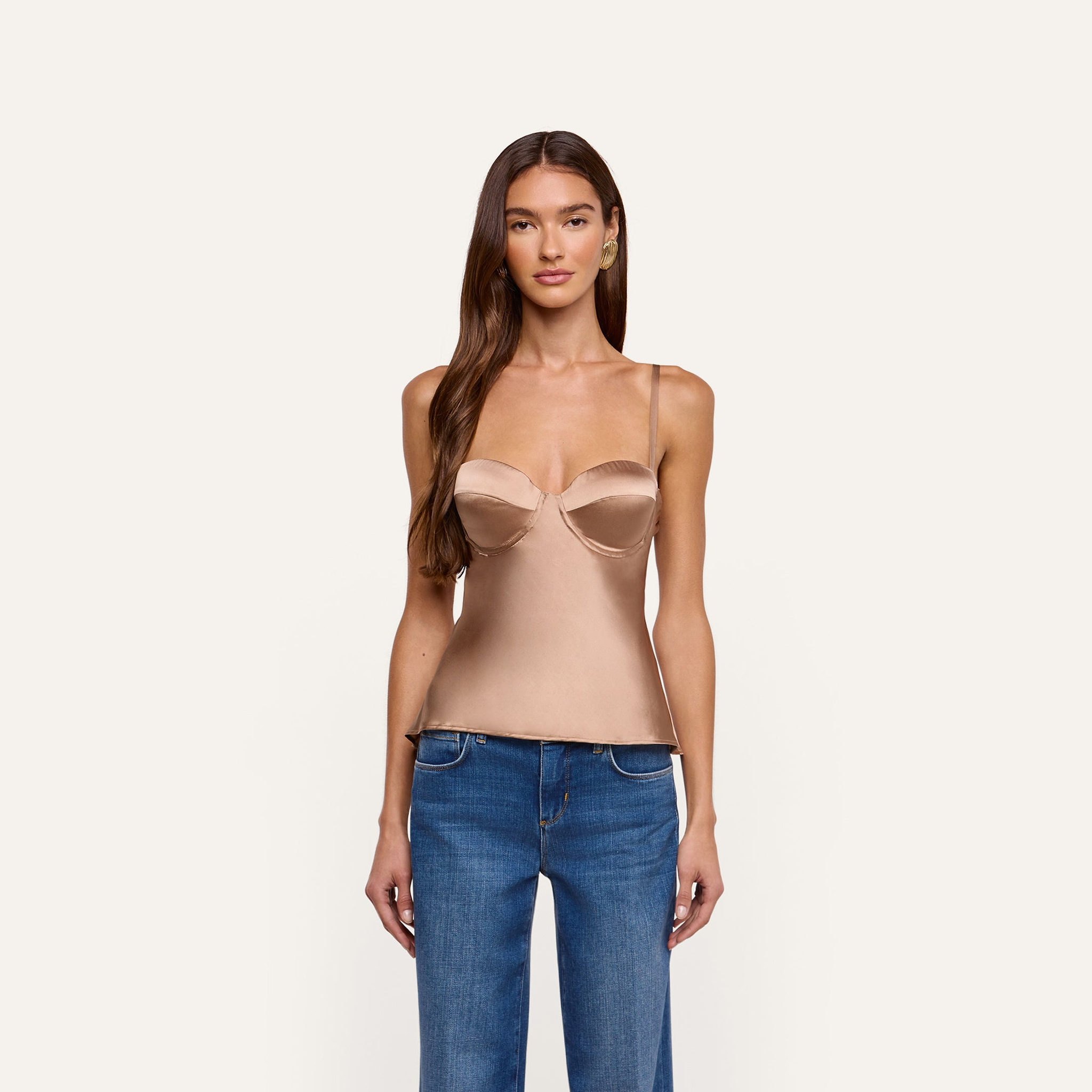 A person wearing an Orlina Silk Camisole, a beige, silk charmeuse corset-style top with thin straps, paired with blue jeans.