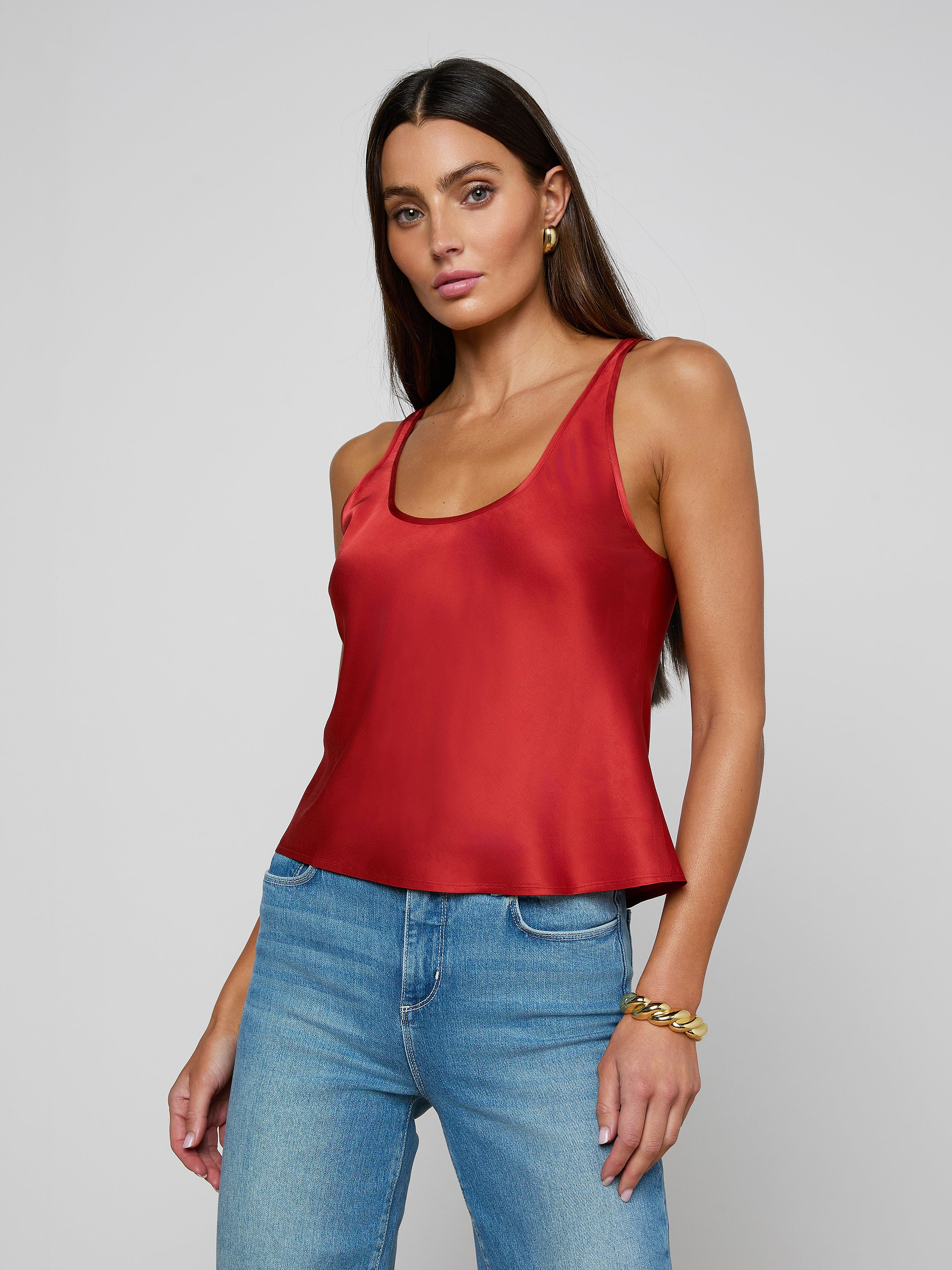 A woman with long dark hair wears the Eddie Tank, a bias-cut satin poppy red sleeveless top with a scoop neckline, paired with blue jeans and accessorized with gold hoop earrings and a gold bracelet, standing against a plain light background.