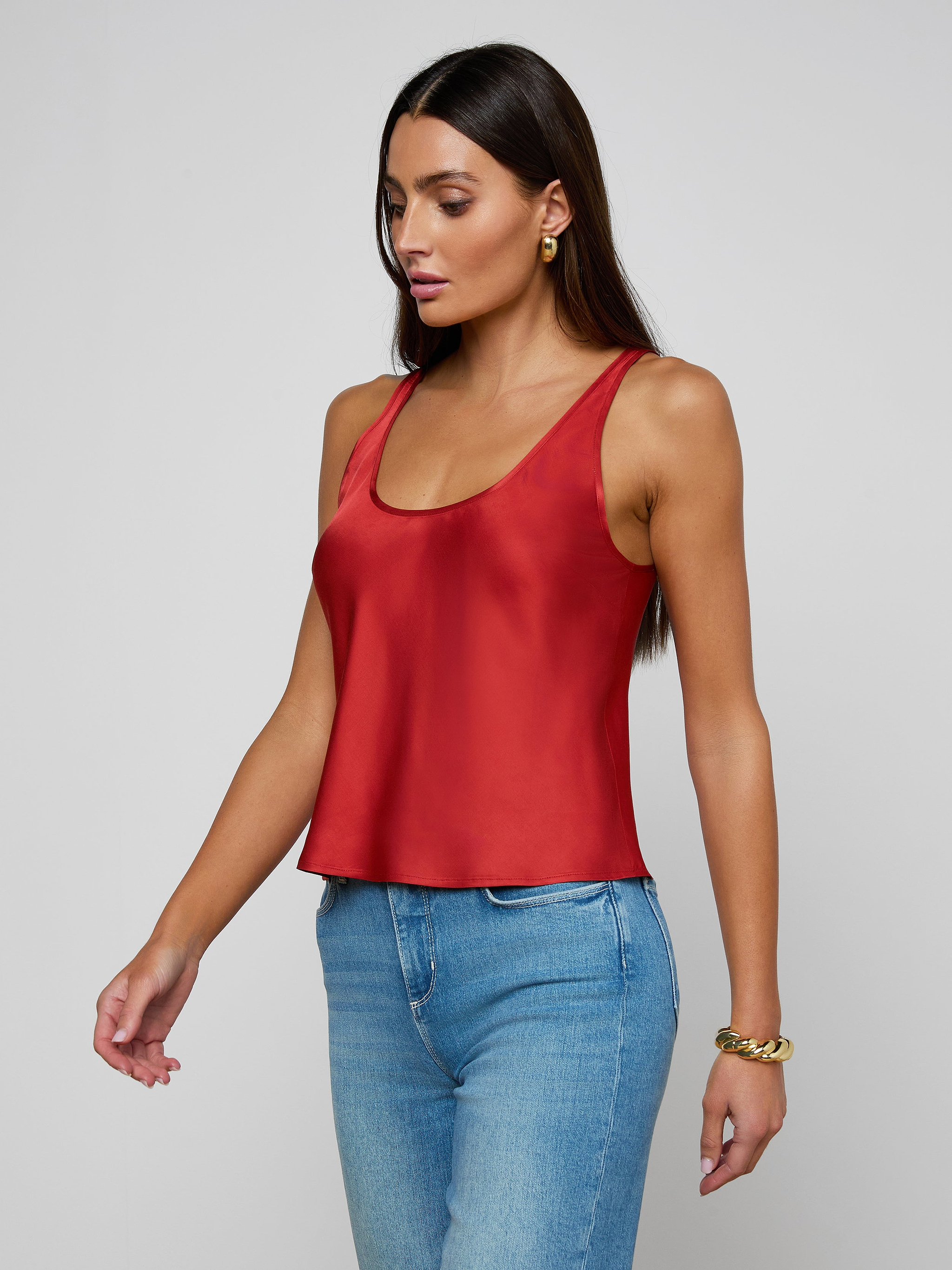 A woman with long brown hair wears the Eddie Tank, a sleeveless poppy red bias-cut satin top with a scoop neckline, paired with blue jeans. She accessorizes with gold hoop earrings and a gold bracelet, standing against a plain light background.