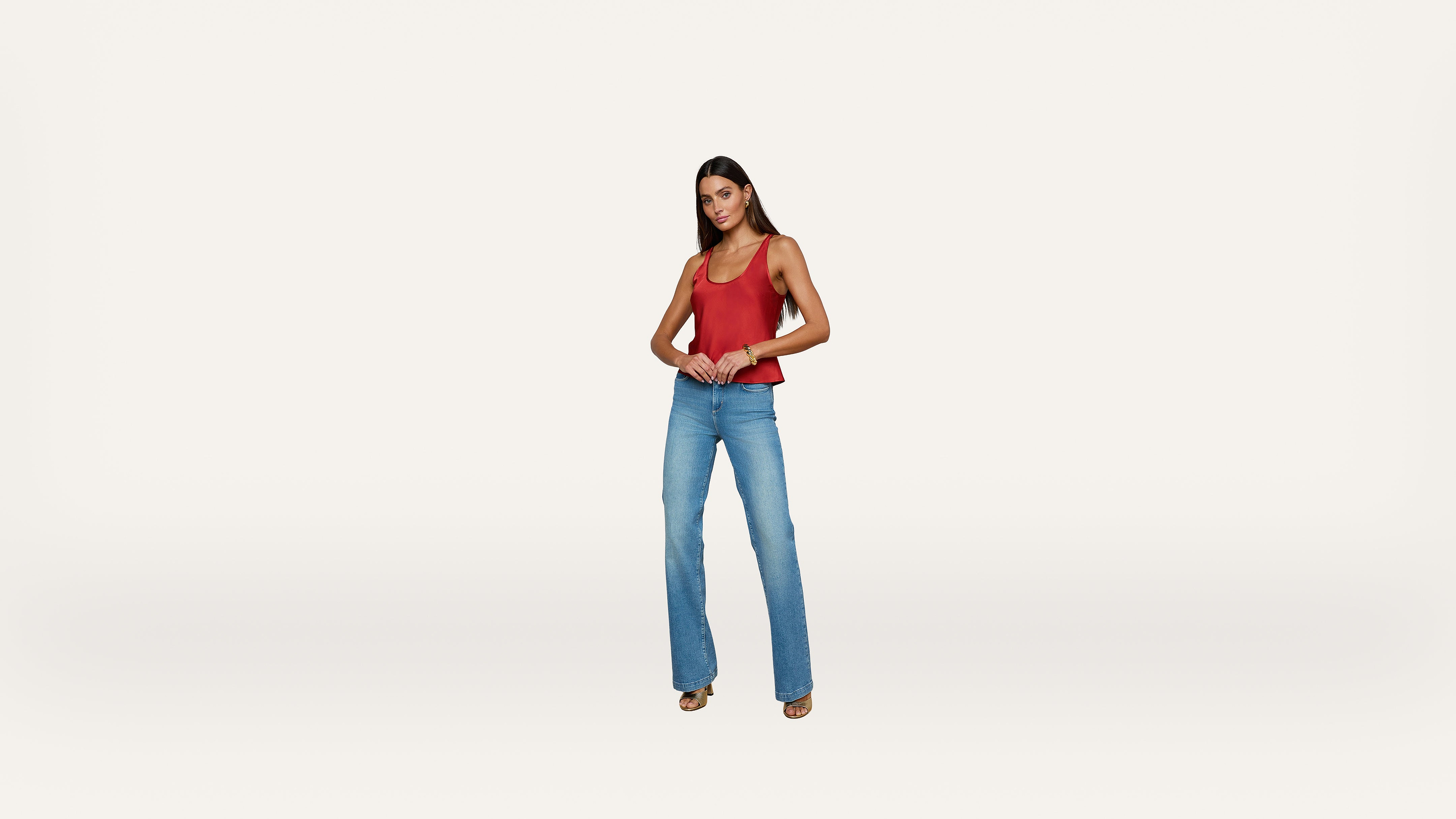 Woman standing against a plain white background, wearing a sleeveless red top, blue flared jeans, and sandals. She has long dark hair and is looking forward with her hands clasped in front.