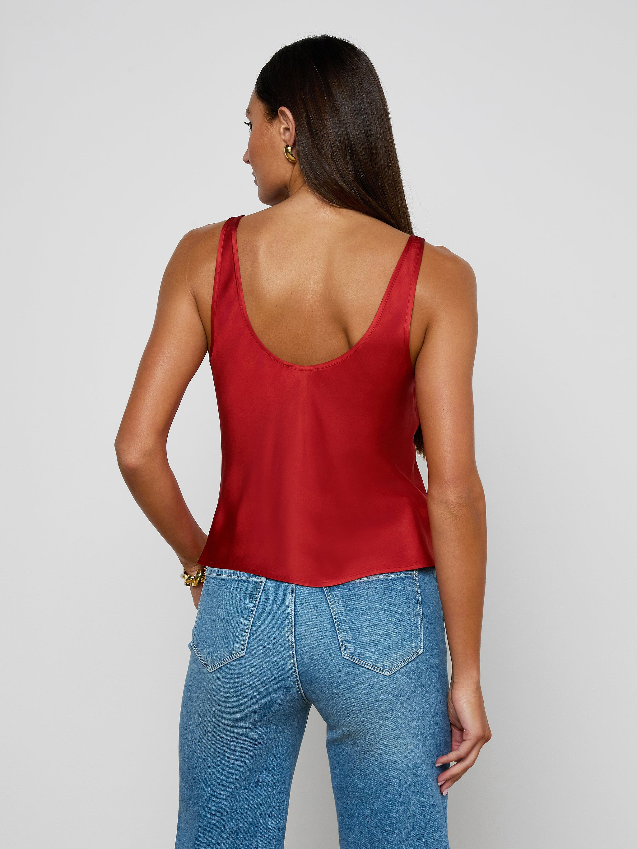 A woman with long brown hair, wearing the Eddie Tank—a bias-cut satin top in poppy red—paired with blue jeans, stands facing away from the camera against a plain light background.