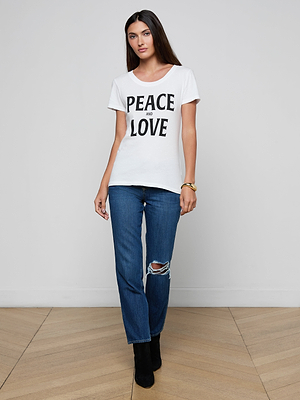 A woman with long brown hair and a neutral expression stands indoors wearing the Cory Graphic Tee, featuring bold PEACE AND LOVE text, paired with blue ripped-knee jeans, black ankle boots, and a gold bracelet.