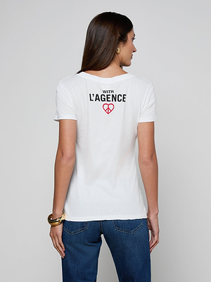 A woman with long brown hair is seen from behind wearing the Cory Graphic Tee in pure white cotton, featuring WITH L’AGENCE and a red peace sign on the upper back, blue jeans, plus gold hoop earrings and bracelets.