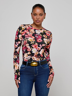 A woman stands against a gray background, wearing the L'AGENCE Tess Long-Sleeve Tee with a colorful floral print, blue jeans, a black belt with a gold buckle, small gold hoop earrings, and her hair pulled back.