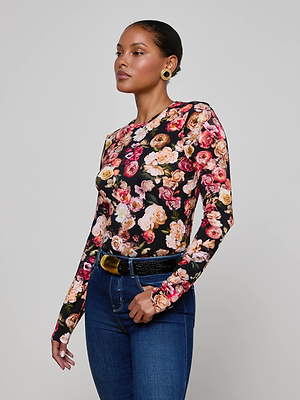 A woman with slicked-back hair wears L'AGENCE's Tess Long-Sleeve Tee in a colorful floral print, gold hoop earrings, blue jeans, and a black belt with a gold buckle, standing against a plain light gray background.