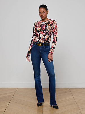 A woman stands indoors against a white wall, wearing the L'AGENCE Tess Long-Sleeve Tee with a floral print, paired with dark blue jeans, a black belt, and black heels. Her hair is pulled back as she looks confidently at the camera.