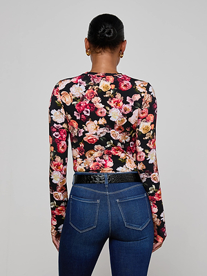 A woman with her hair in a neat bun is seen from behind, wearing the L'AGENCE Tess Long-Sleeve Tee with a colorful floral print and high-waisted blue jeans with a black belt, set against a plain light gray background.