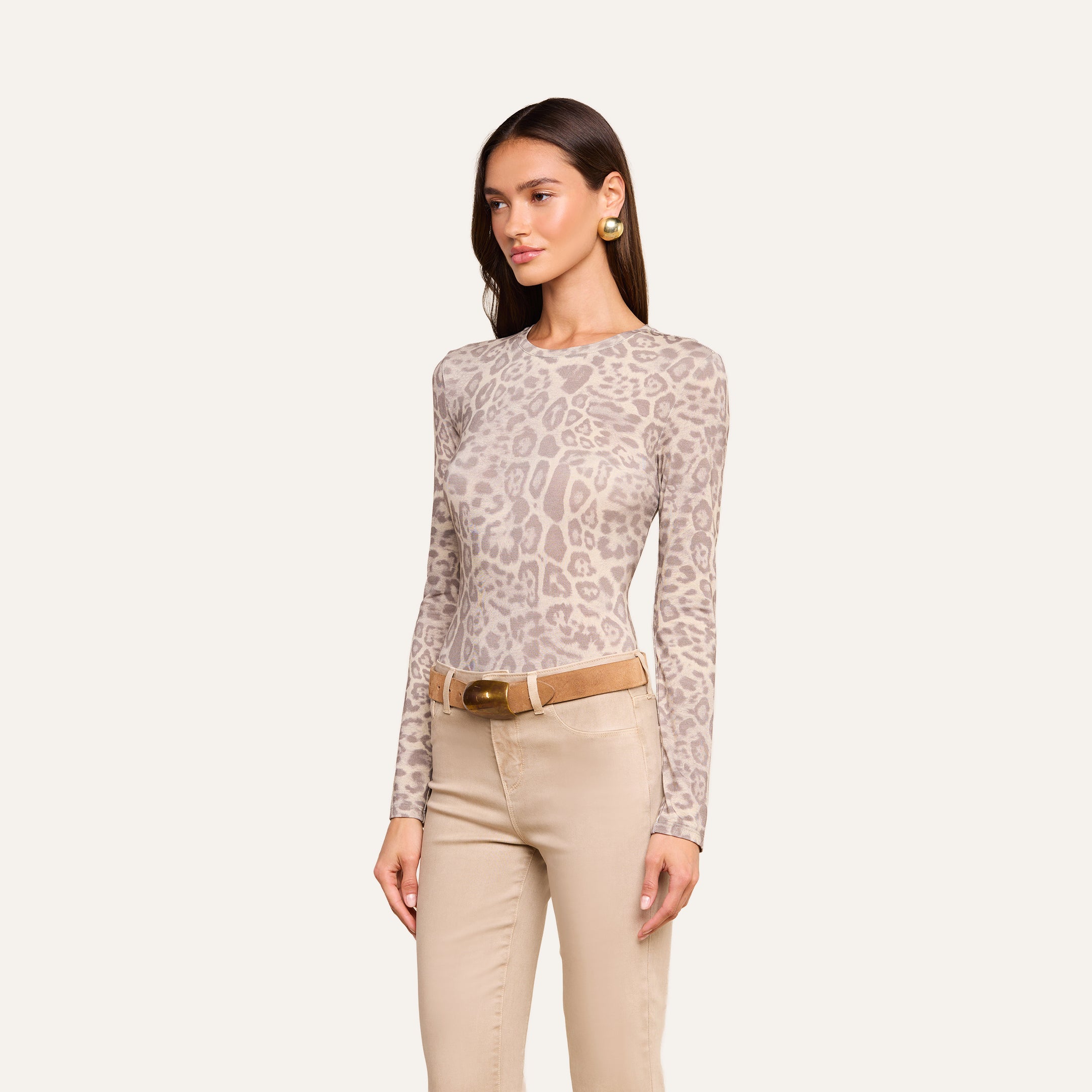 A woman wearing the Tess Long Sleeve Tee in a slim fit grey leopard print, paired with tan pants, a brown belt, and gold earrings, stands against a plain light background.