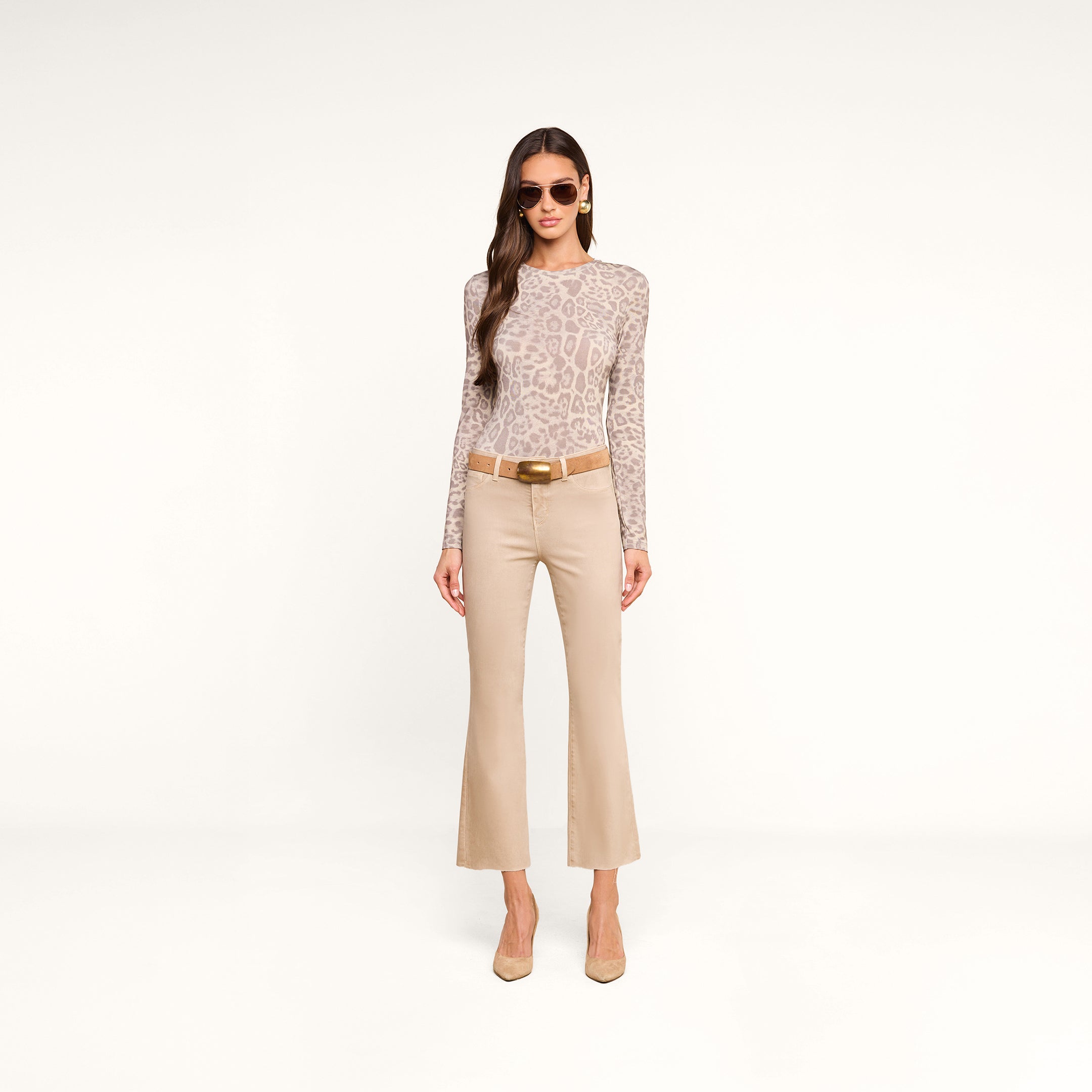 A woman stands against a plain white background wearing sunglasses, the Tess Long Sleeve Tee in a slim fit grey leopard print, tan cropped pants with a brown belt, and beige heels.