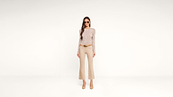 Woman in sunglasses, a long-sleeve patterned top, beige flared pants, and heels stands against a plain white background, facing forward with hands by her sides.