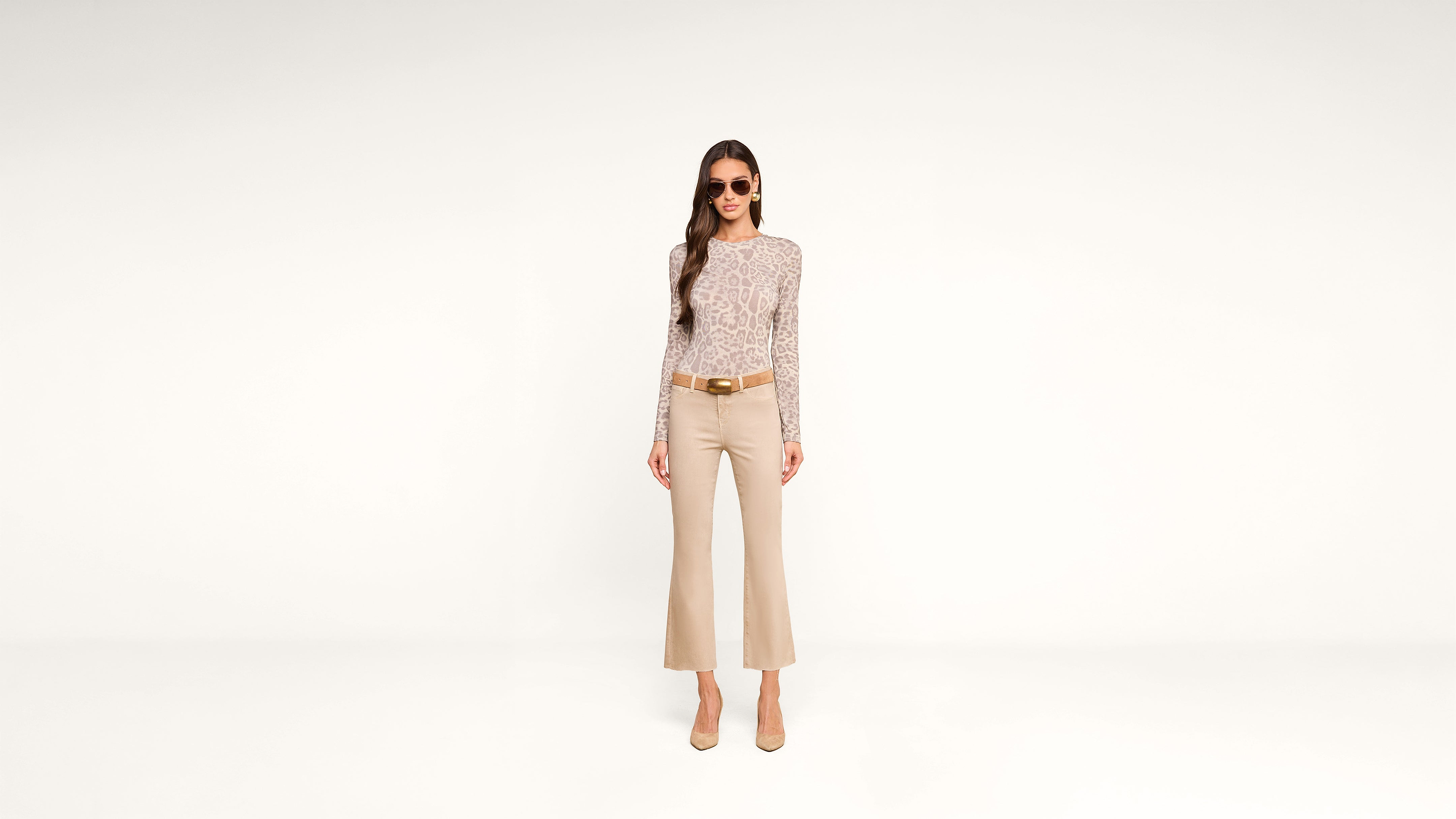 Woman in sunglasses, a long-sleeve patterned top, beige flared pants, and heels stands against a plain white background, facing forward with hands by her sides.
