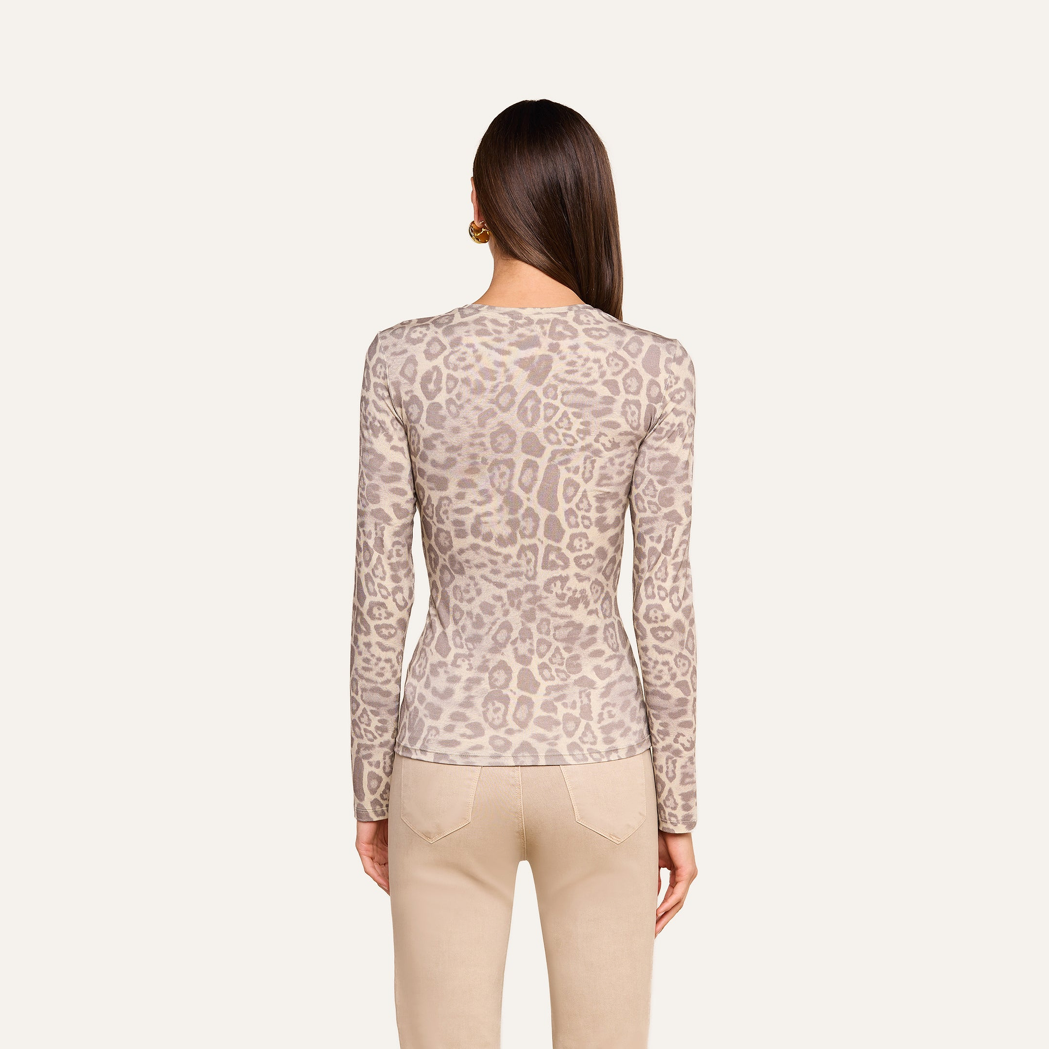 A woman with straight brown hair is shown from the back against a plain light background wearing the Tess Long Sleeve Tee—a slim fit, long-sleeve grey leopard print top—paired with beige pants.