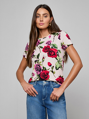 A woman with long brown hair wears the Ressi Fitted Tee, a bestselling white short-sleeved slim-cut top with a red and pink rose print, paired with blue jeans. She stands with one hand in her pocket against a plain gray background.