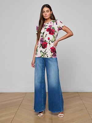 A woman with long brown hair and a neutral expression poses indoors wearing the Ressi Fitted Tee in white with a red and pink floral print, wide-leg blue jeans, and white open-toe heels, with one hand on her hip.