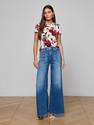 A woman with long brown hair stands indoors on a wooden floor, wearing the Ressi Fitted Tee featuring red and pink roses, wide-leg blue jeans, and white open-toe heels. She has a neutral expression.