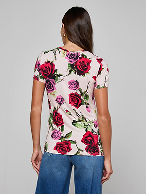 A woman with long brown hair, wearing the bestselling Ressi Fitted Tee—slim cut, short-sleeved, white with a red and pink rose print—and blue jeans stands facing away from the camera against a plain background.