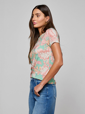 A woman with long brown hair stands in profile, wearing the Ressi Fitted Tee—a slim cut, pink and green floral print short-sleeve top—and blue jeans against a plain light gray background.