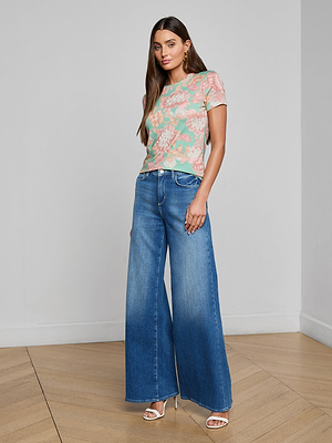 A woman stands on a wooden floor wearing the bestselling Ressi Fitted Tee in a pastel floral print, wide-leg blue jeans, and white heeled sandals, posing against a plain light-colored wall.