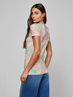 A woman with long brown hair stands sideways, looking over her shoulder. She wears the Ressi Fitted Tee in a pastel floral print and blue jeans against a plain light gray background.