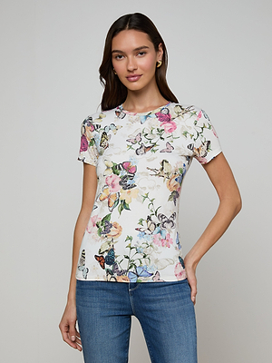A woman stands against a plain background, wearing our Ressi Fitted Tee in stretch jersey with a colorful floral and butterfly print. She pairs it with blue jeans, her long brown hair and a slight smile completing the look.