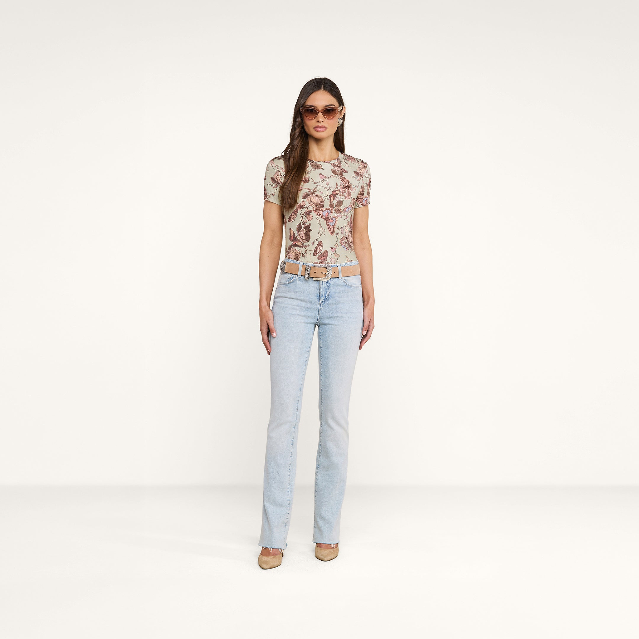 A woman stands facing forward against a plain white background, wearing sunglasses, heels, a belt, light blue jeans, and the Ressi Fitted Tee—a slim cut short-sleeve patterned top.