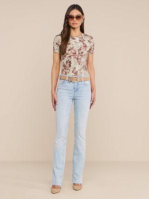 A woman stands against a plain beige background wearing a bestselling Ressi Fitted Tee with butterfly print, light blue high-waisted jeans, a beige belt, nude heels, and sunglasses.