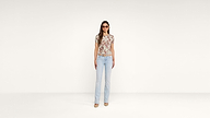 A woman stands against a plain white background wearing light blue jeans, a floral short-sleeve top, beige heels, and sunglasses.