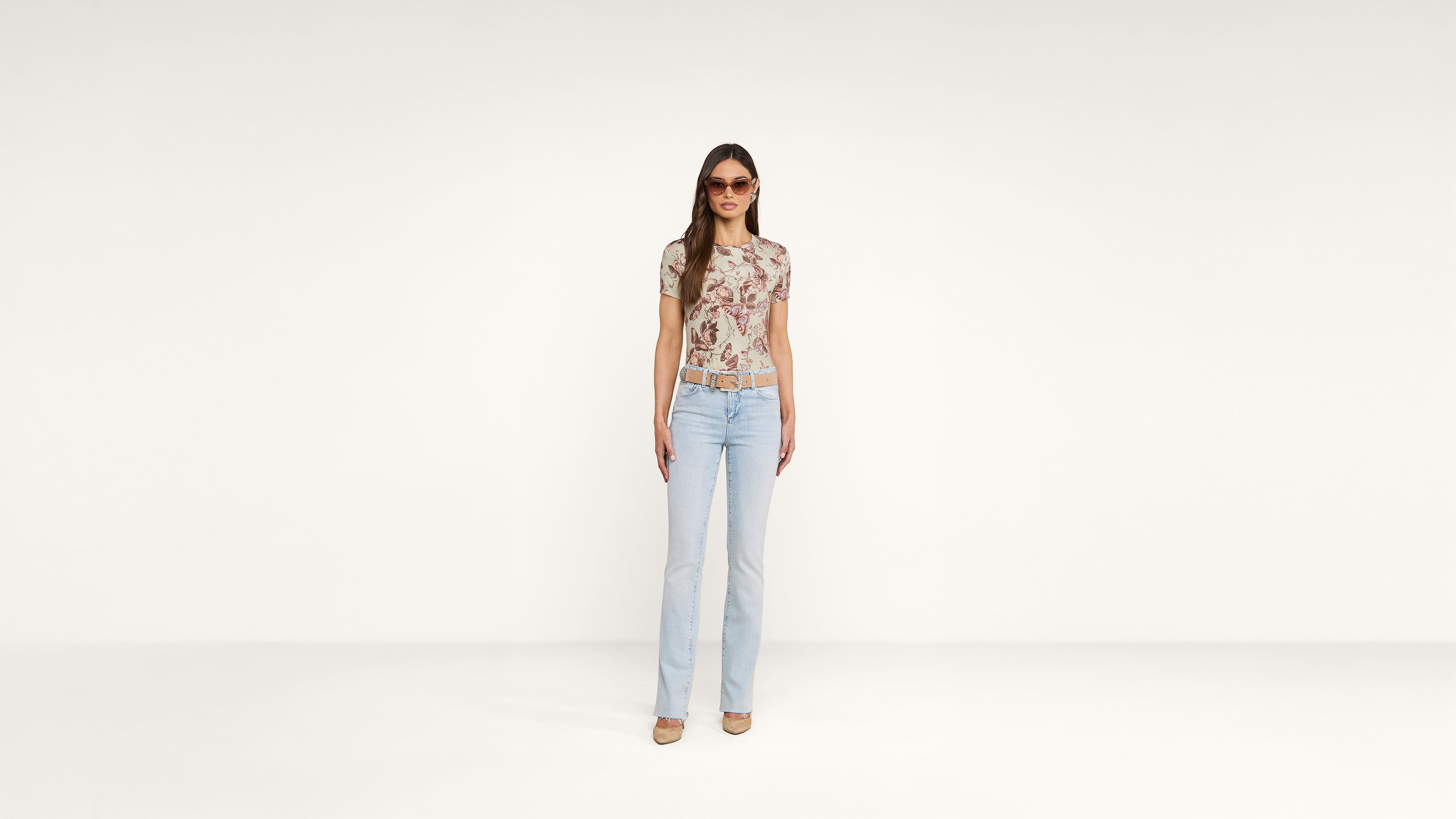 A woman stands against a plain white background wearing light blue jeans, a floral short-sleeve top, beige heels, and sunglasses.