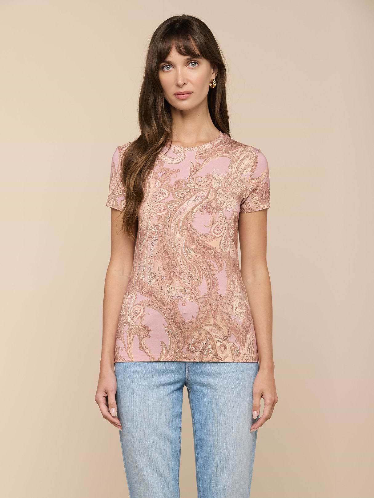 Una mujer de pelo castaño largo lleva la camiseta Ressi Fitted Tee, un éxito de ventas, con estampado paisley rosa y beige, de manga corta y corte entallado, combinada con unos vaqueros azul claro, sobre un fondo beige.