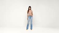 Woman with long brown hair stands against a plain white background, wearing a short-sleeved peach top, light blue flared jeans, and brown shoes, with one hand in her pocket.