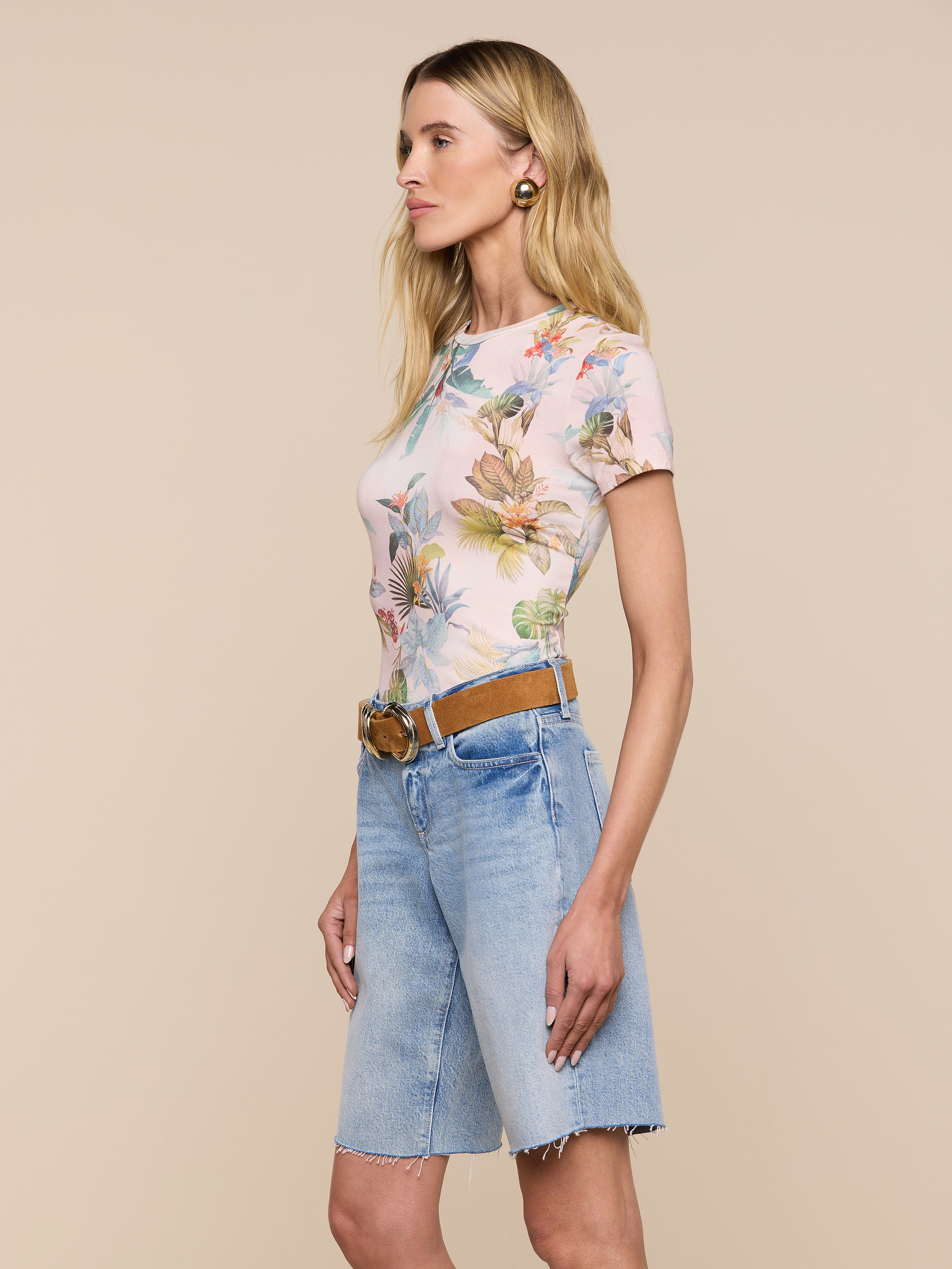 Woman standing in profile against a light background, wearing the Ressi Fitted Tee—a slim-cut pastel floral short-sleeve top—tucked into knee-length denim shorts with a tan belt and gold hoop earrings.