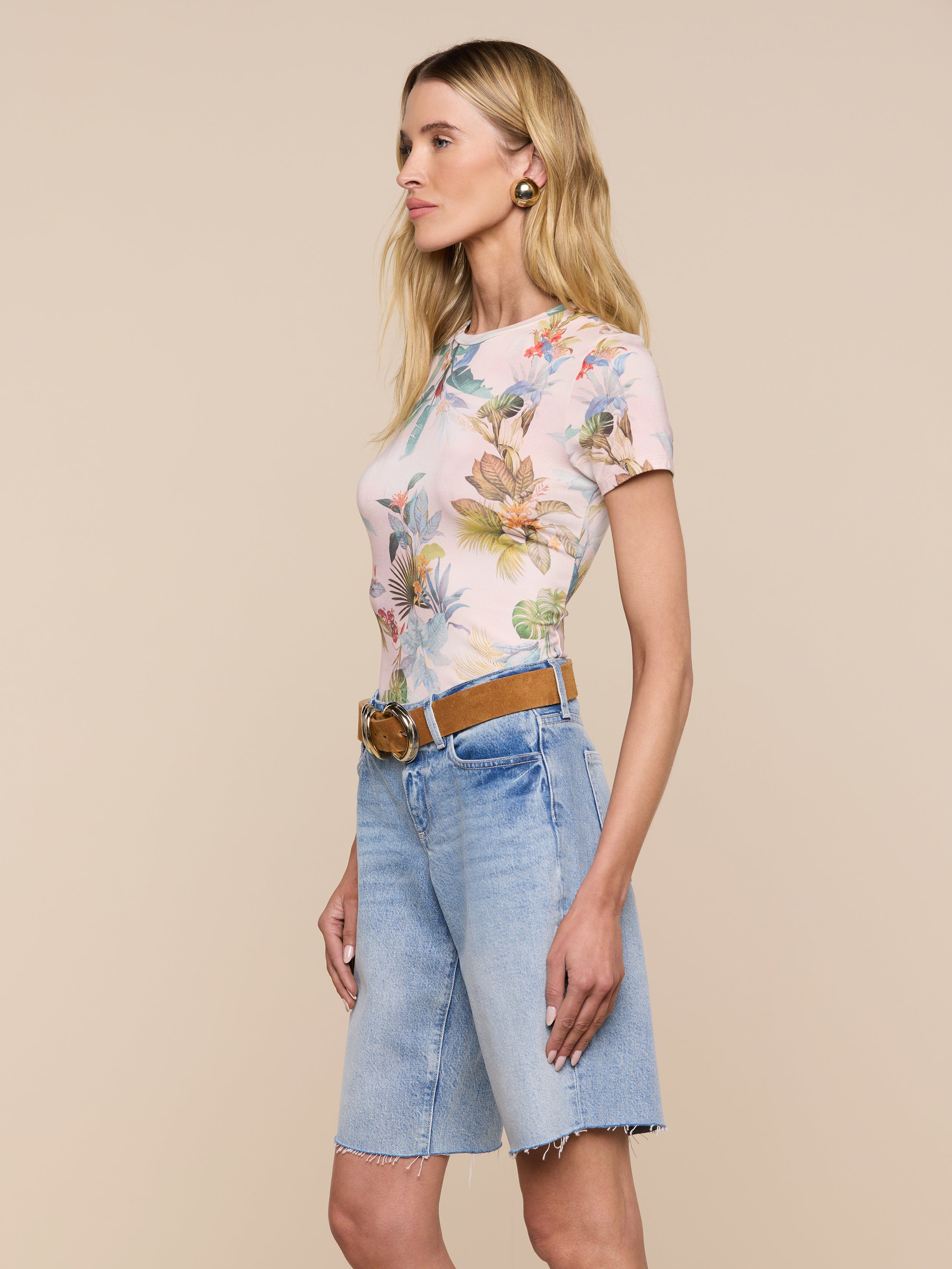 Woman standing in profile against a light background, wearing the Ressi Fitted Tee—a slim-cut pastel floral short-sleeve top—tucked into knee-length denim shorts with a tan belt and gold hoop earrings.