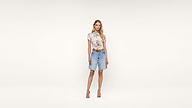 A woman stands against a plain white background wearing a floral short-sleeve top, blue denim shorts, a brown belt, and beige heels.