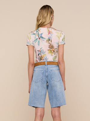 A woman with long, wavy blonde hair is seen from behind in a pastel palm leaf print Ressi Fitted Tee and high-waisted light blue raw-hem denim Bermuda shorts, standing against a beige background.