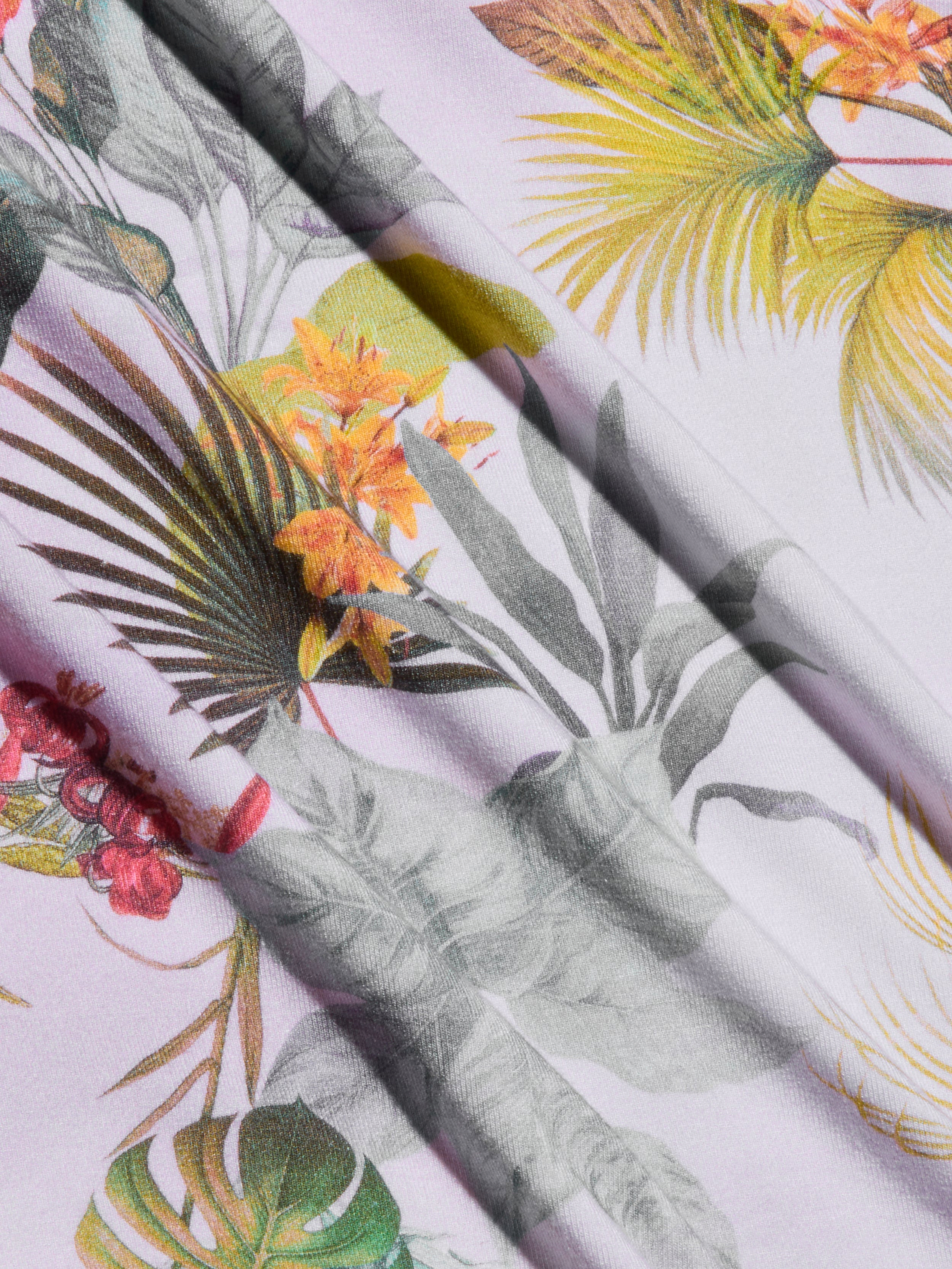 Close-up of the Ressi Fitted Tee, showcasing fabric with a palm leaf print and tropical floral pattern—featuring green palm leaves, orange and yellow flowers, plus hints of red and gray on a light background. The tee’s fabric displays soft folds and gentle shadows.