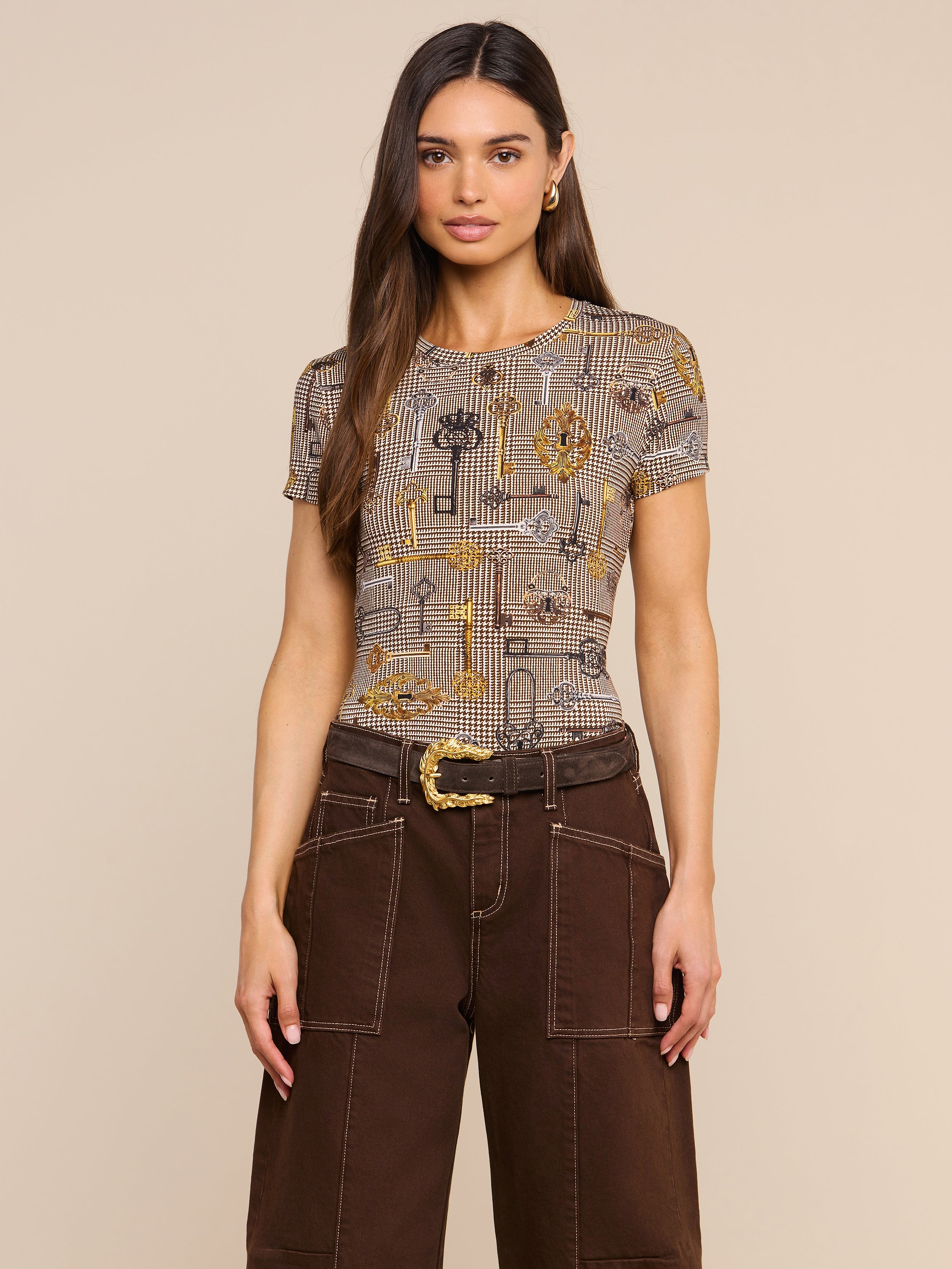 Wearing the bestselling Ressi Fitted Tee with a brown houndstooth pattern, a woman with long brown hair pairs it with wide-leg brown pants featuring white stitching and a belted waist, standing against a plain light background.