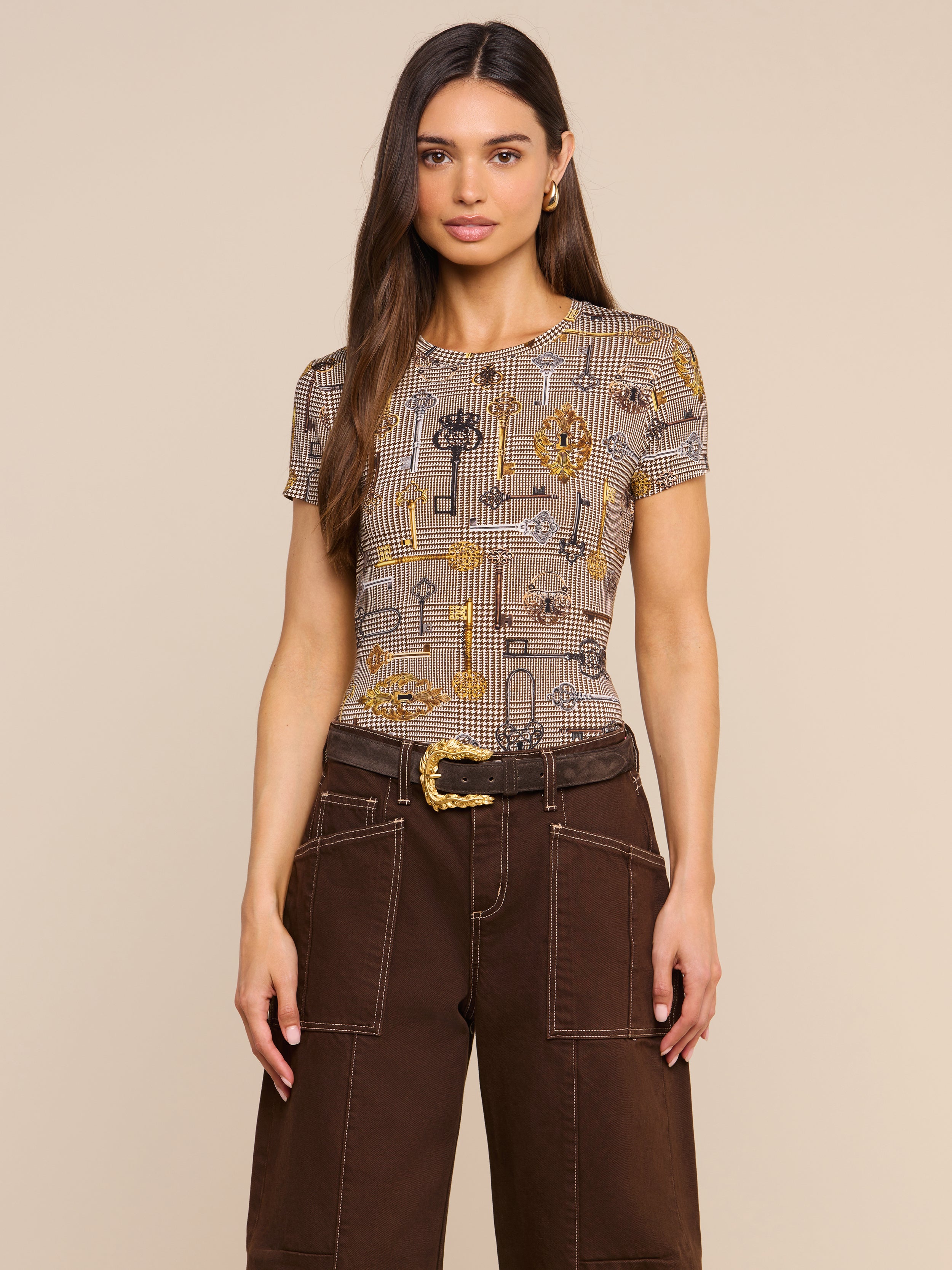 Wearing the bestselling Ressi Fitted Tee with a brown houndstooth pattern, a woman with long brown hair pairs it with wide-leg brown pants featuring white stitching and a belted waist, standing against a plain light background.