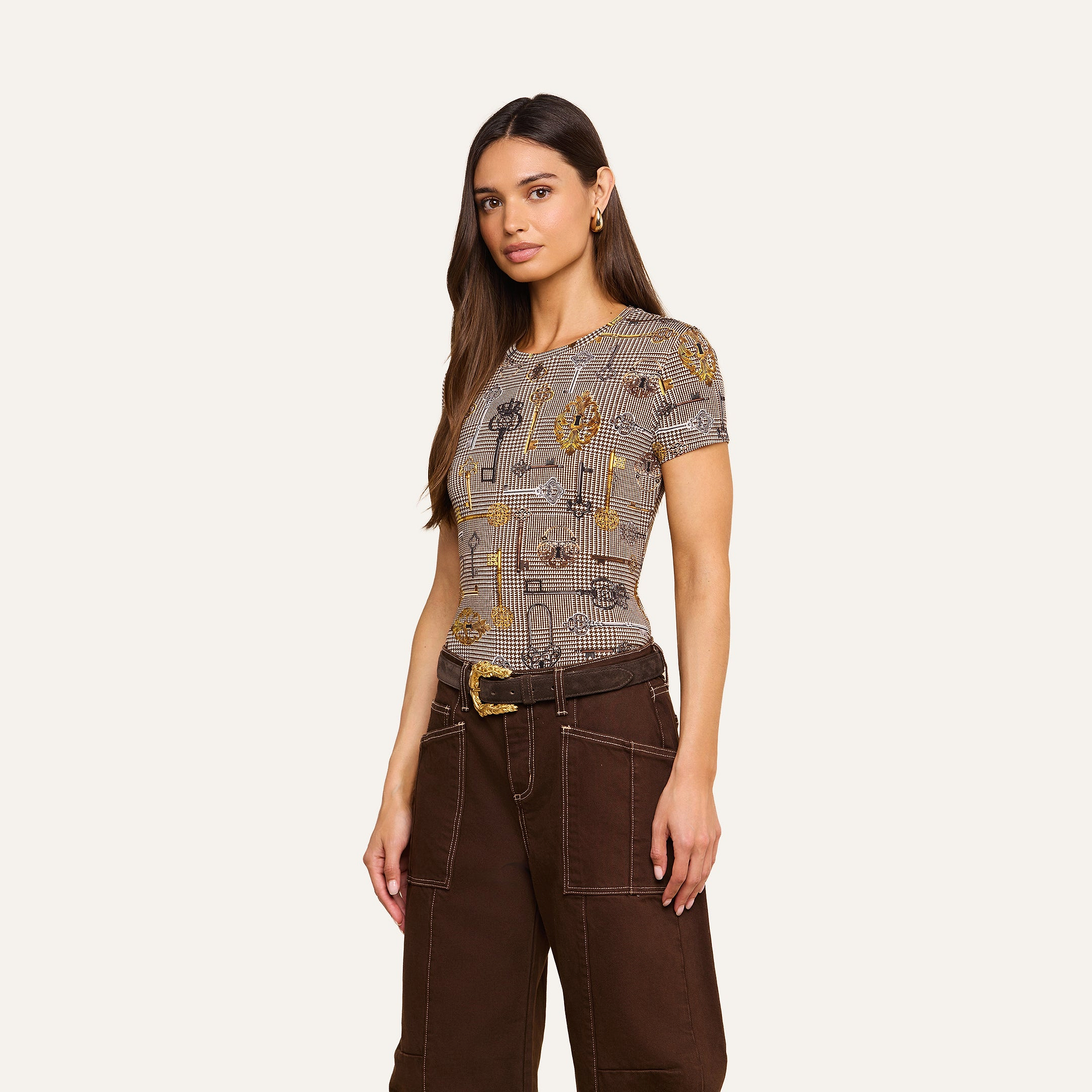 A woman with long brown hair wears the bestselling Ressi Fitted Tee and high-waisted dark brown pants with white stitching, standing against a plain light background.