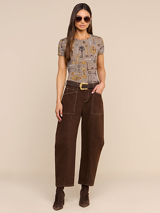 A woman wearing sunglasses, the Ressi Fitted Tee—a soft printed jersey short-sleeve top—loose brown pants with white stitching, a textured belt, and brown boots stands against a beige background with one hand in her pocket.