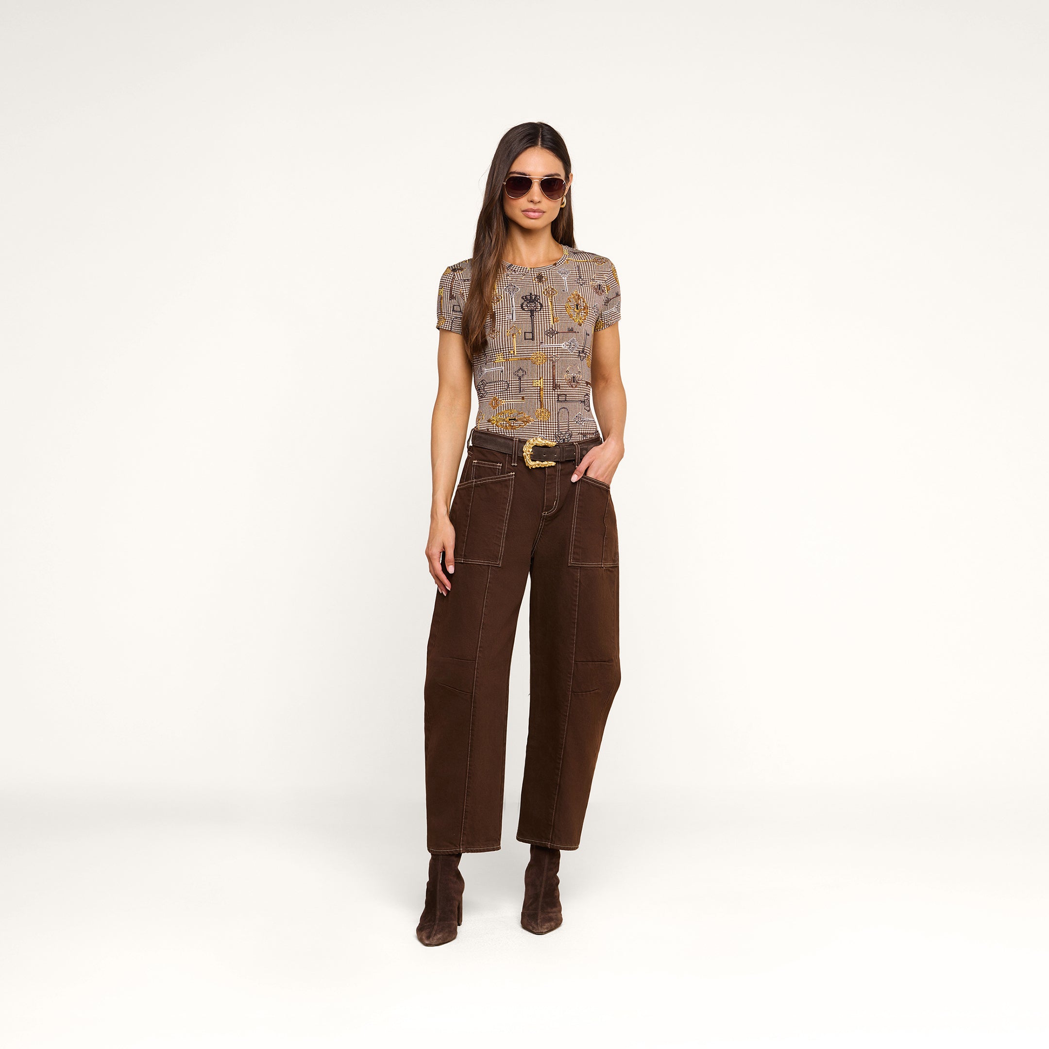 A woman wearing sunglasses, the Ressi Fitted Tee with short sleeves, slim cut brown pants with patch pockets, and brown boots stands against a plain white background with one hand in her pocket.