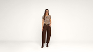 A woman wearing sunglasses, a short-sleeve patterned top, brown pants, and brown boots stands against a plain white background with one hand in her pocket.