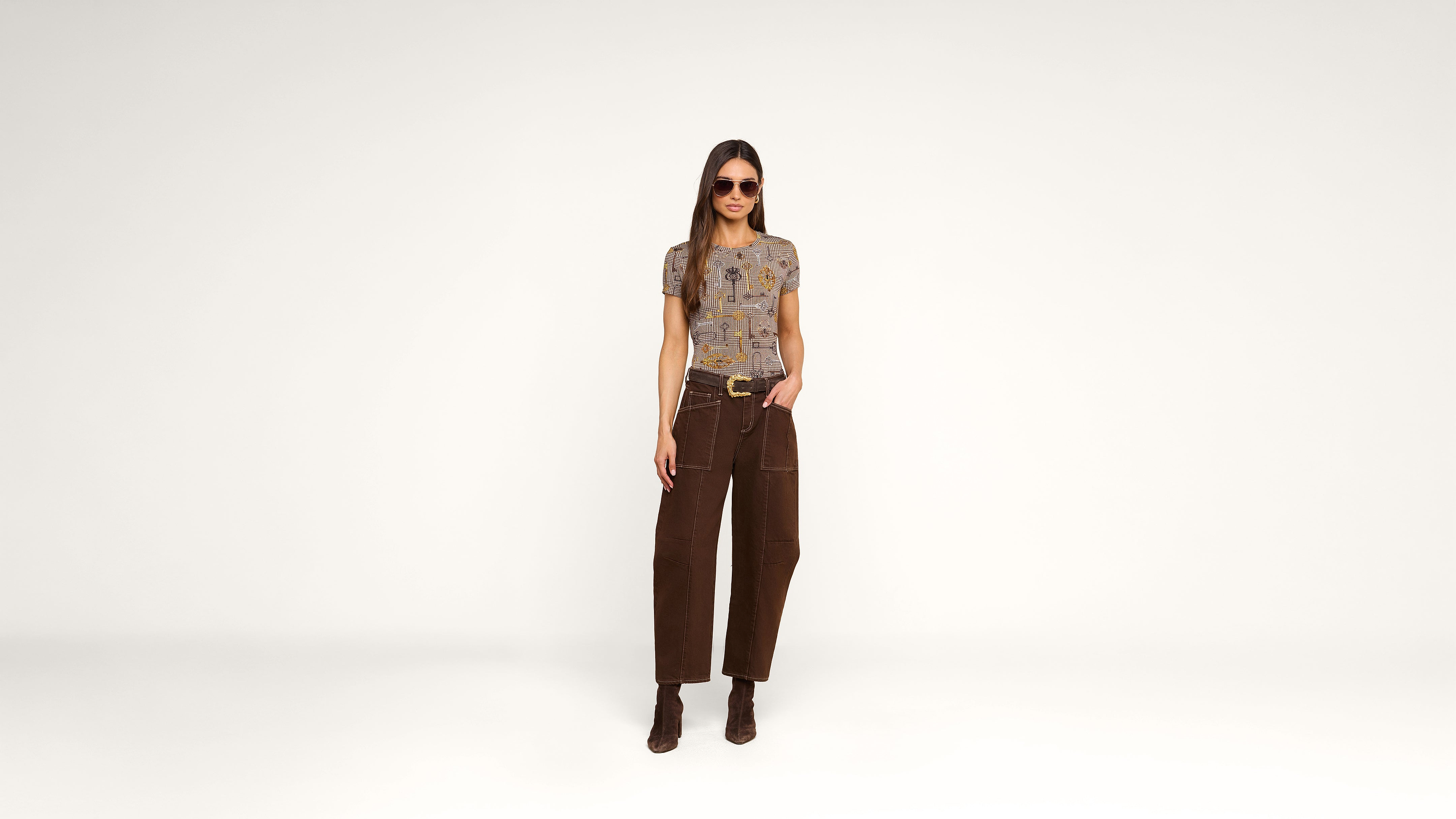 A woman wearing sunglasses, a short-sleeve patterned top, brown pants, and brown boots stands against a plain white background with one hand in her pocket.