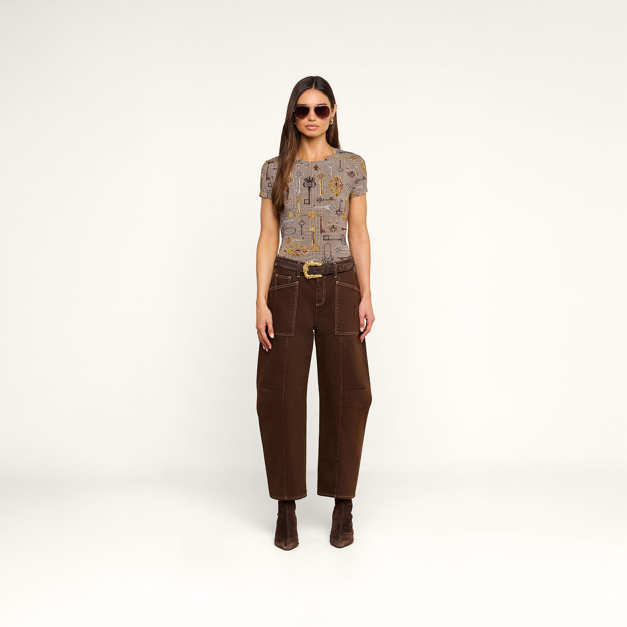 A woman wearing sunglasses, the bestselling Ressi Fitted Tee with a patterned short-sleeve design, brown high-waisted wide-leg pants, and brown boots stands facing forward against a plain white background.