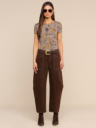 Woman stands against a beige background wearing sunglasses, the Ressi Fitted Tee in a brown houndstooth pattern, high-waisted brown pants with large pockets and a belt, and brown ankle boots.