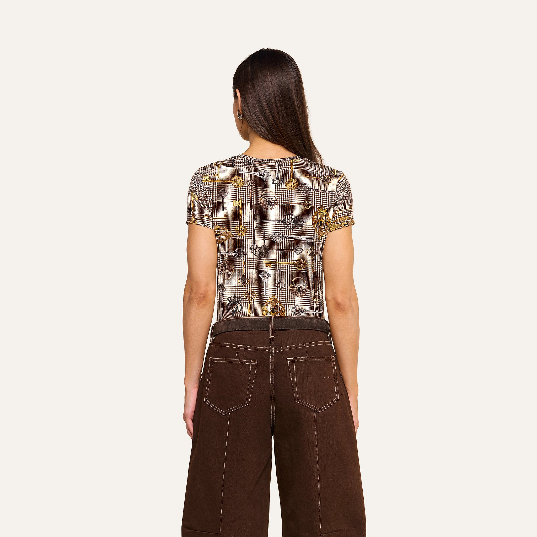 A woman with long brown hair stands facing away, wearing the bestselling Ressi Fitted Tee and high-waisted brown pants with white stitching, against a plain light background.
