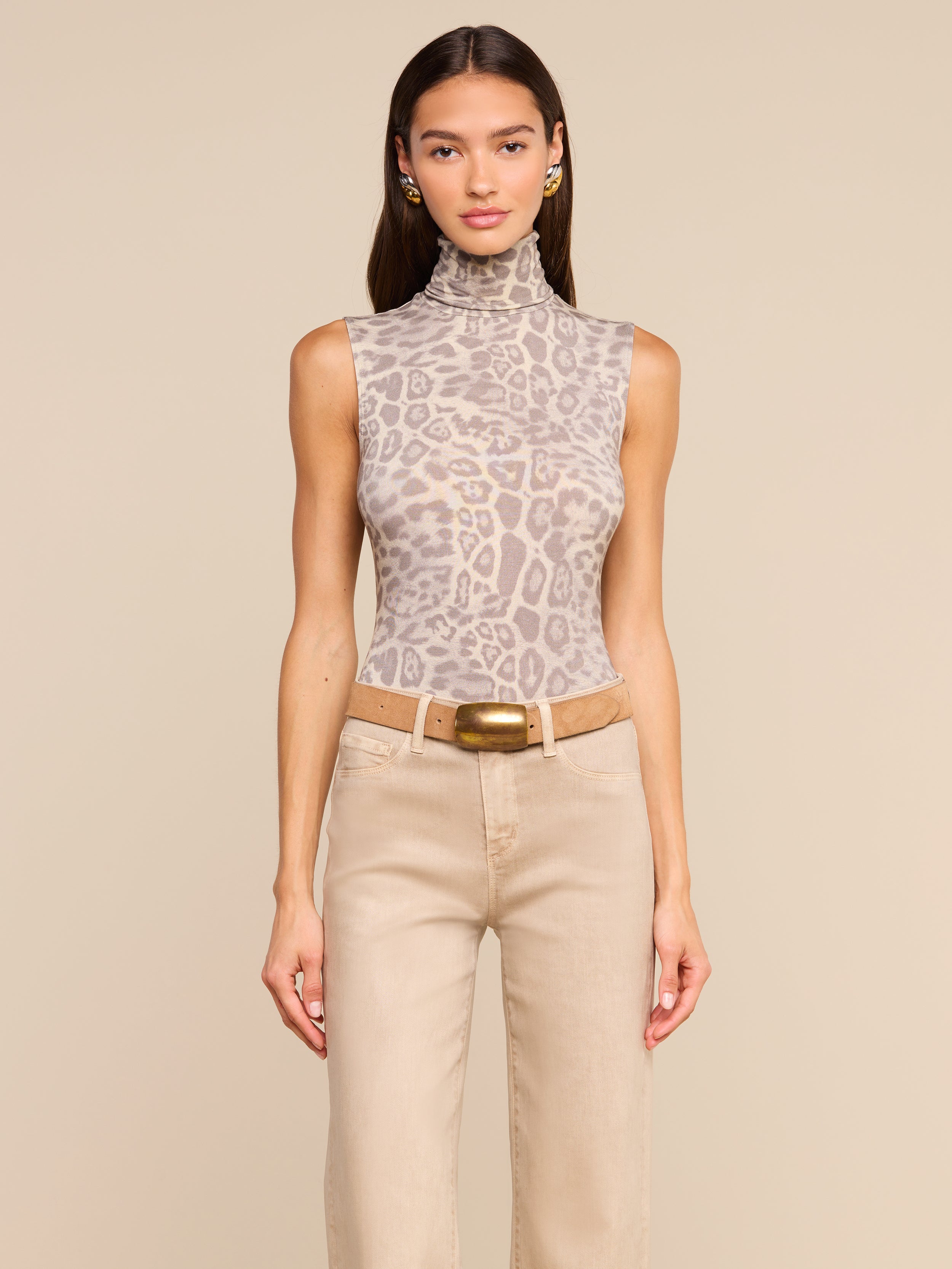 A woman stands facing forward, wearing the Ceci Sleeveless Turtleneck in a grey leopard print, paired with beige pants, a wide belt with a gold buckle, and small hoop earrings. She has long straight hair and a neutral expression.